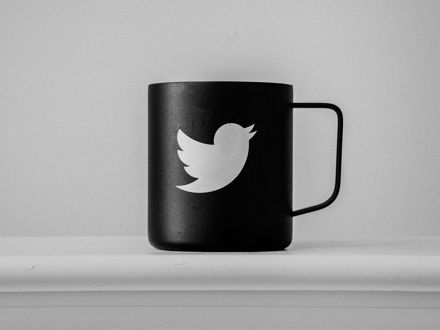 A black mug with a white Twitter bird logo on it. The mug is on a white surface in front of a white wall.