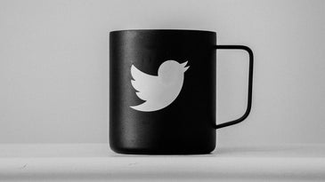 A black mug with a white Twitter bird logo on it. The mug is on a white surface in front of a white wall.