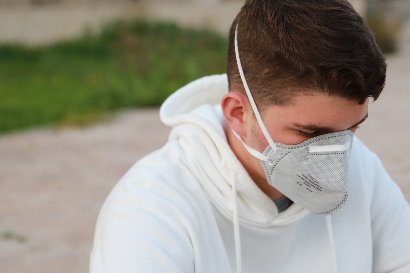 A long COVID patient with short brown hair and a white sweatshirt wearing a respirator mask while sitting on the street