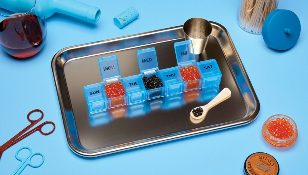 Metal tray wth vials of blue and orange pulls surrounded by medical tools and a glass on red wine on a light blue background