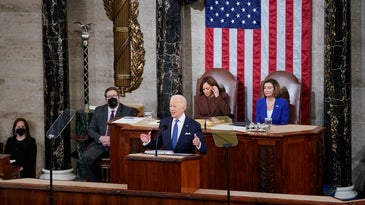Biden State of the Union 2022 with the US president, vice president, and speaker of the House of Representatives in front of an American flag and two other individuals wearing COVID masks in the Capitol