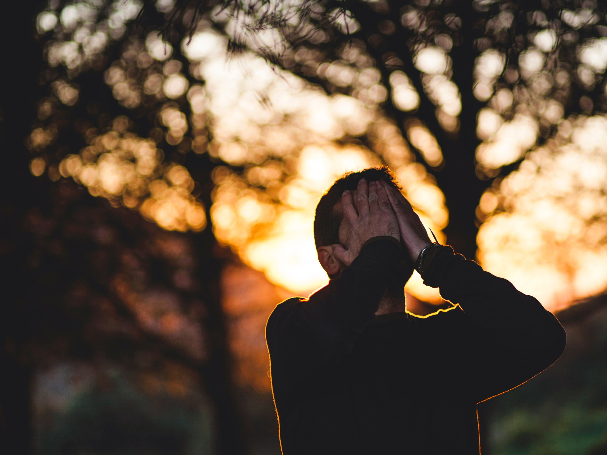 Een man die bij zonsondergang onder bomen staat en zijn gezicht in zijn handen houdt omdat hij gestrest of angstig is.