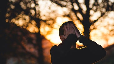 A man standing under some trees outside at sunset, holding his face in his hands because he is stressed out or anxious.
