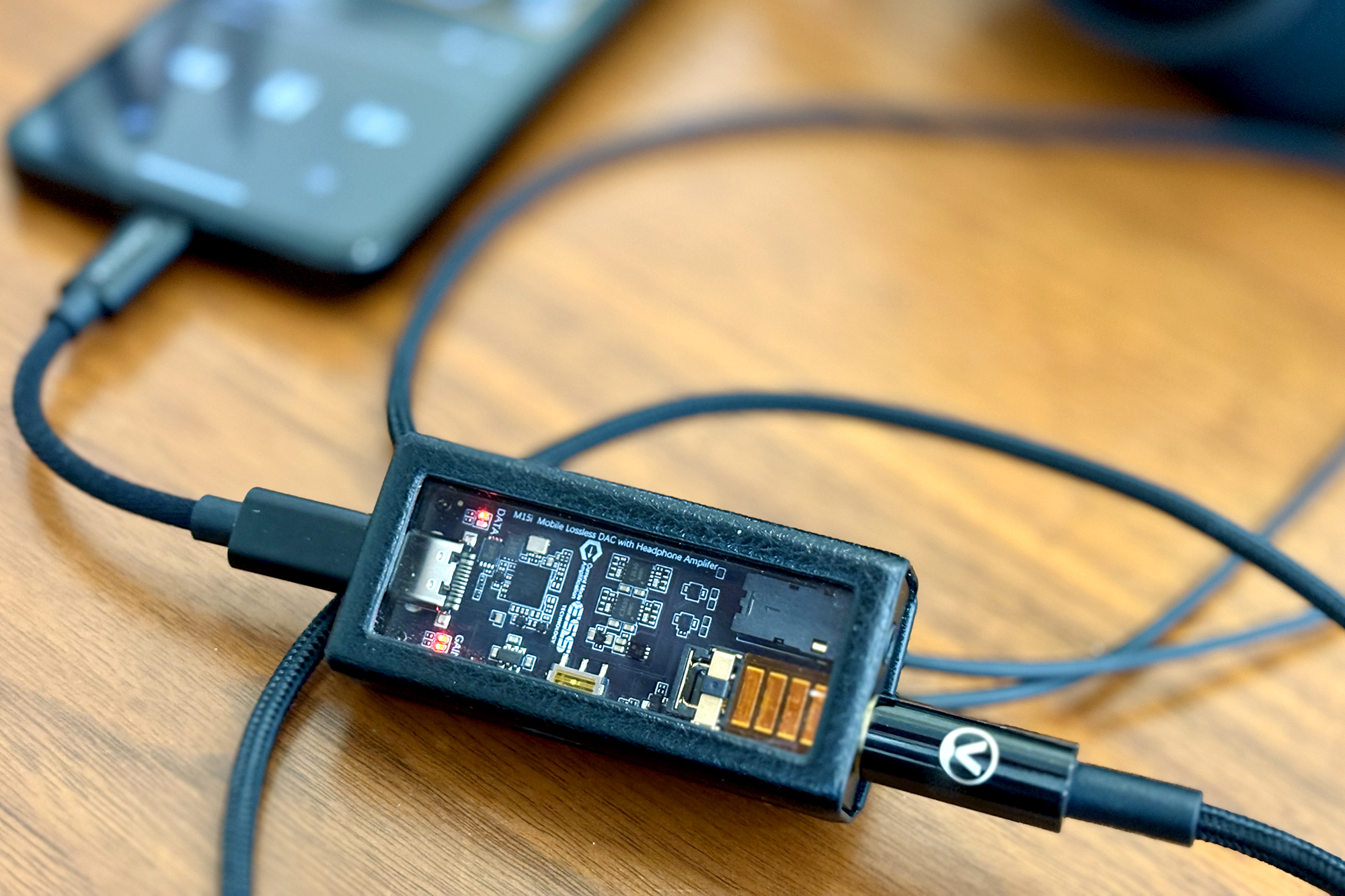 Rectangular Questyle M15i USB DAC dongle in a black leather slipcase on a wood table.