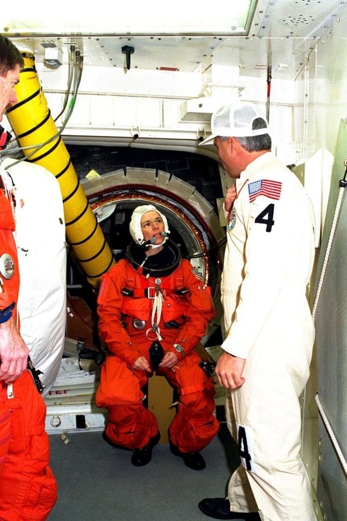 Female astronaut in a bulky orange spacesuit on a space shuttle talking to NASA techs in white form-fitting suits