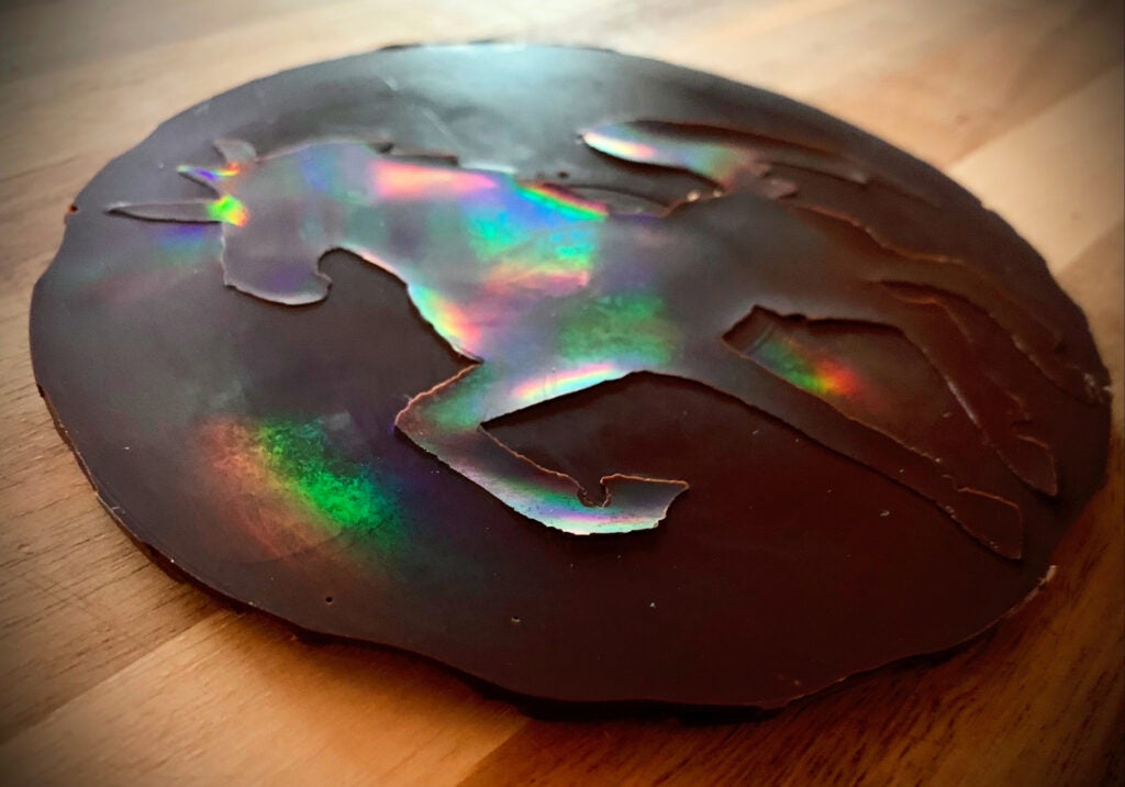 A disk of chocolate with a unicorn standing in shallow relief on its surface. Both the disk and the unicorn are reflecting rainbow colors.