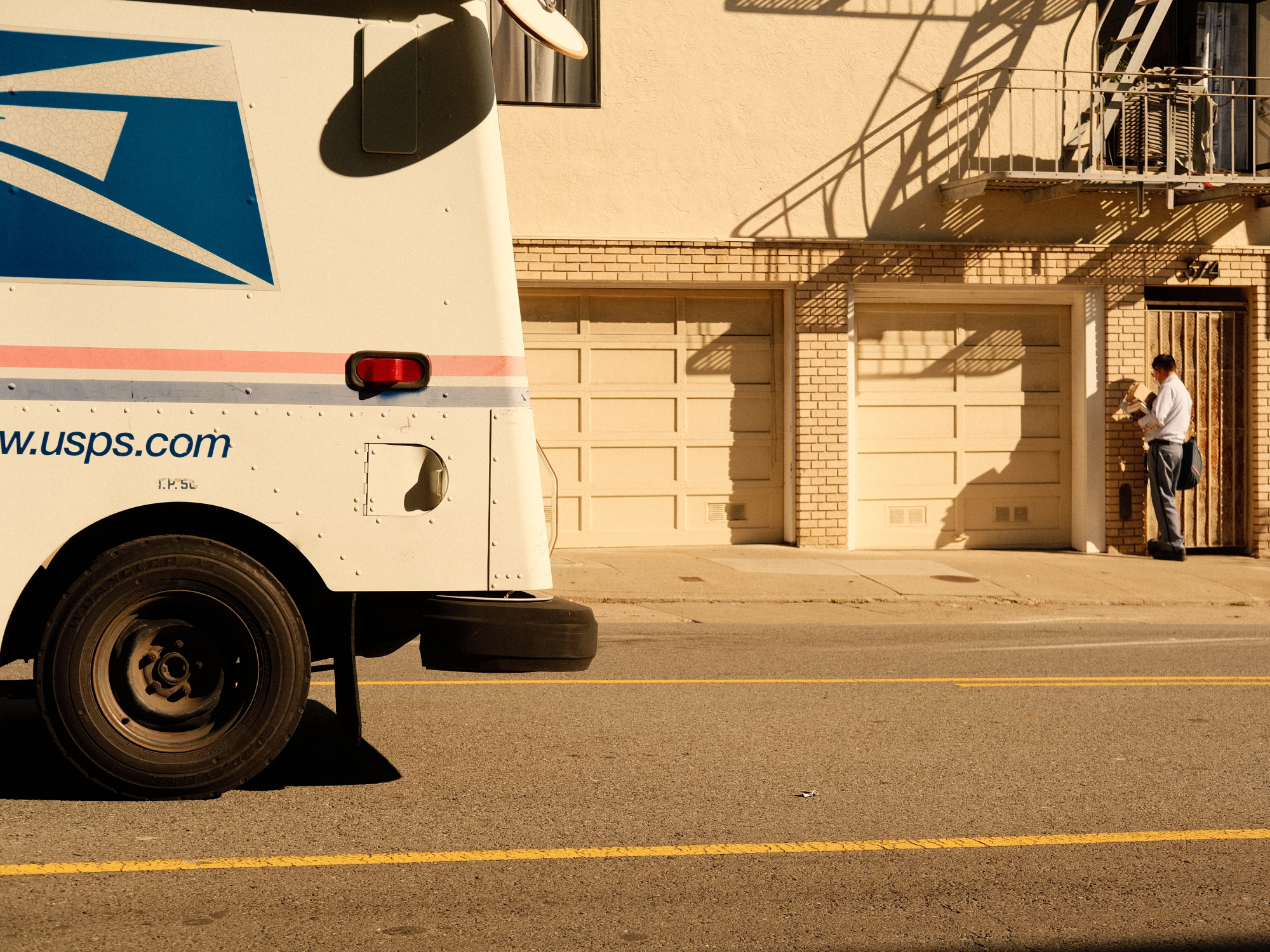 Een Amerikaanse postbeambte bezorgt pakketten aan een bruin woongebouw, met een USPS-vrachtwagen geparkeerd op straat op de voorgrond.