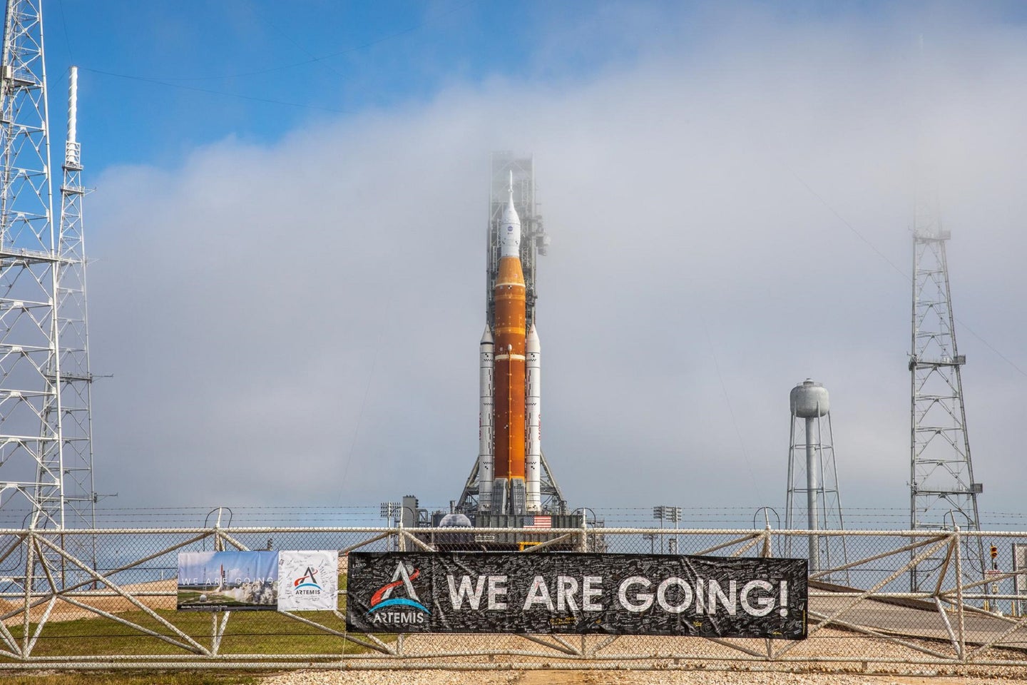 Orange NASA SLS rocket at launchpad at Kennedy Space Center in Florida with a brown banner that says "we are going"