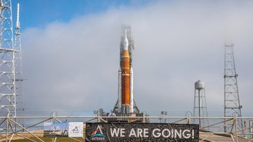 Orange NASA SLS rocket at launchpad at Kennedy Space Center in Florida with a brown banner that says 