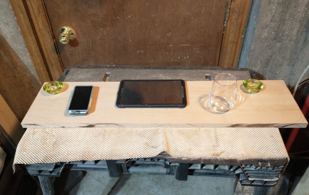 A piece of wood with a phone, tablet, wine glass, and candle on it, to set up a layout for a DIY bathtub tray.