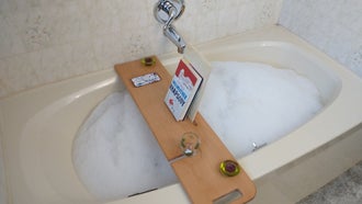 A bathtub full of bubbles with a homemade wooden bath tray on it, complete with a book, a phone, a candle holder, and a wine glass holder.