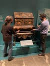 Two art conservators holding a tall painted wooden cabinet from both sides in a blue-walled museum in New York City
