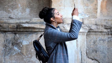 A woman standing in front of a stone wall and looking through her phone at something off-screen, maybe scanning text on a sign.