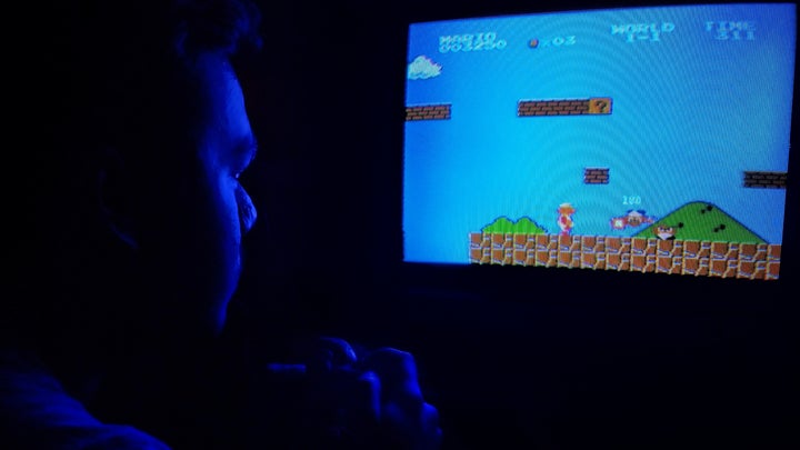 A man sitting in a dark room, his face illuminated only by the blue glow of a cathode-ray TV as he plays a retro Nintendo game.