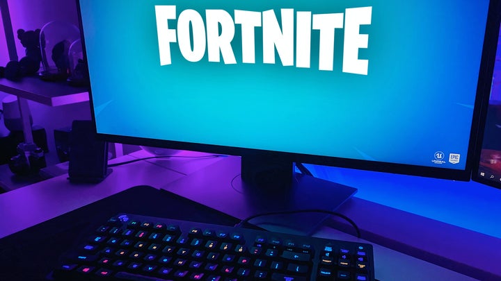 A computer with the title of the video game Fortnite on its screen, in a room lit by blue and purple lights.