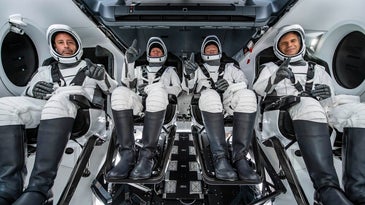 four men sit in spacesuits on a spacecraft