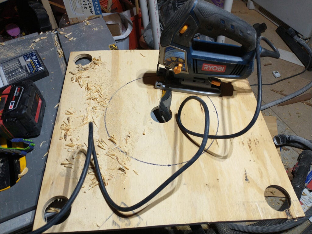 The plywood top of a DIY shop vac dust separator, showing the pipe holes in each corner and a jigsaw in the process of cutting out the center bucket hole.