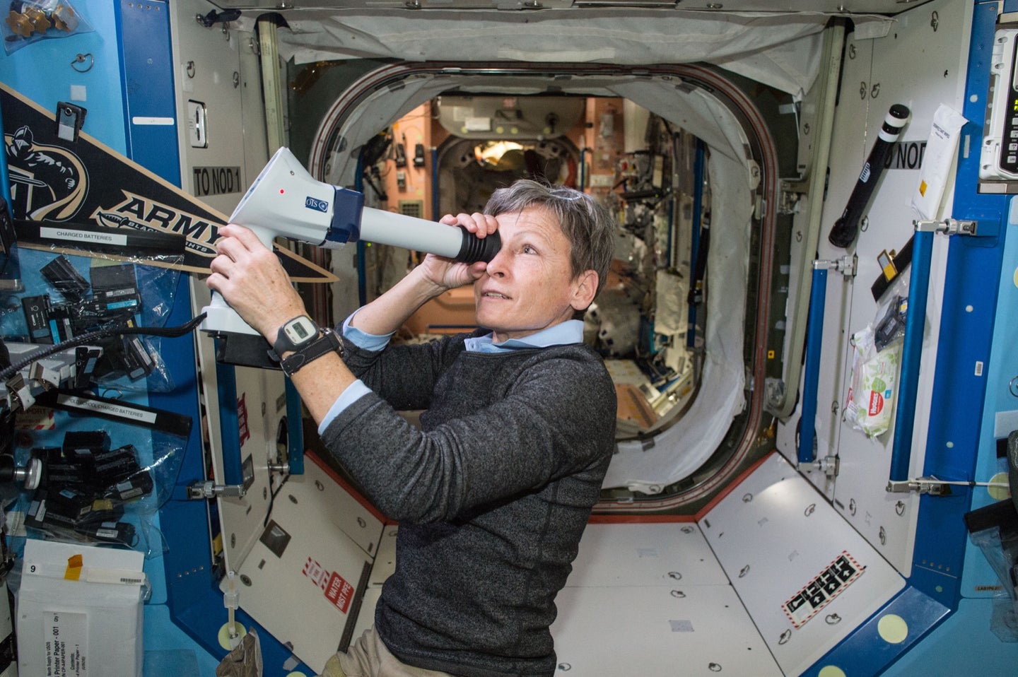 a woman astronaut with short hair and glasses propped on her head looks through a long tube with a lens and camera which takes pictures of her eye. she's aboard the international space station
