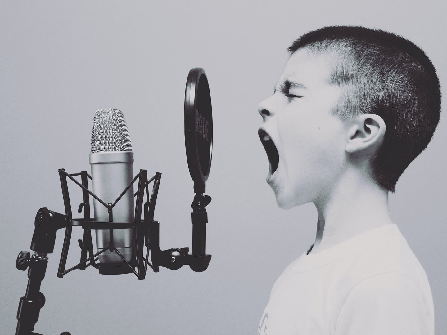 A child with a shaved head and white shirt shouting or singing into a mice with a pop filter in front of it.