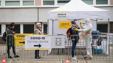 people wait in a line outside a white tent to get tested for covid by a medical practitioner