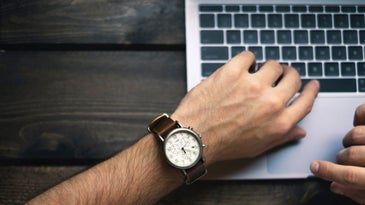 A person using a silver Macbook laptop on a gray wooden surface, while looking at a watch on their wrist.