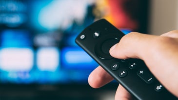 A close-up of a hand holding a TV remote with an out-of-focus TV screen in the background.
