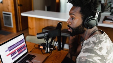 A young Black man wearing headphones and speaking into a microphone while sitting at a wooden table and using a laptop.