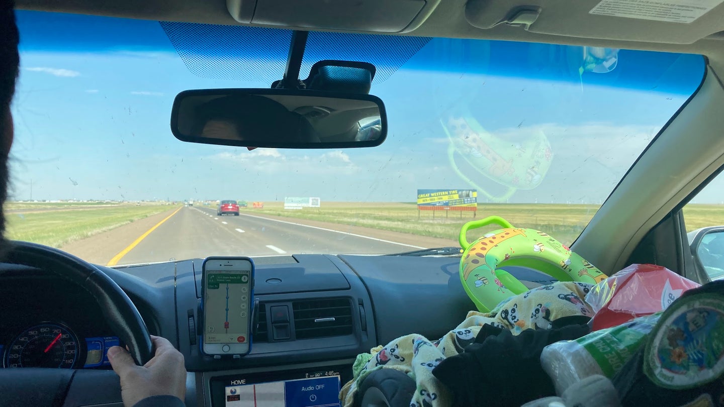 Looking through the windshield of a car driving down a flat road between open fields under blue skies, with one person in the driver's seat and a toddler in a carseat in the passenger seat.
