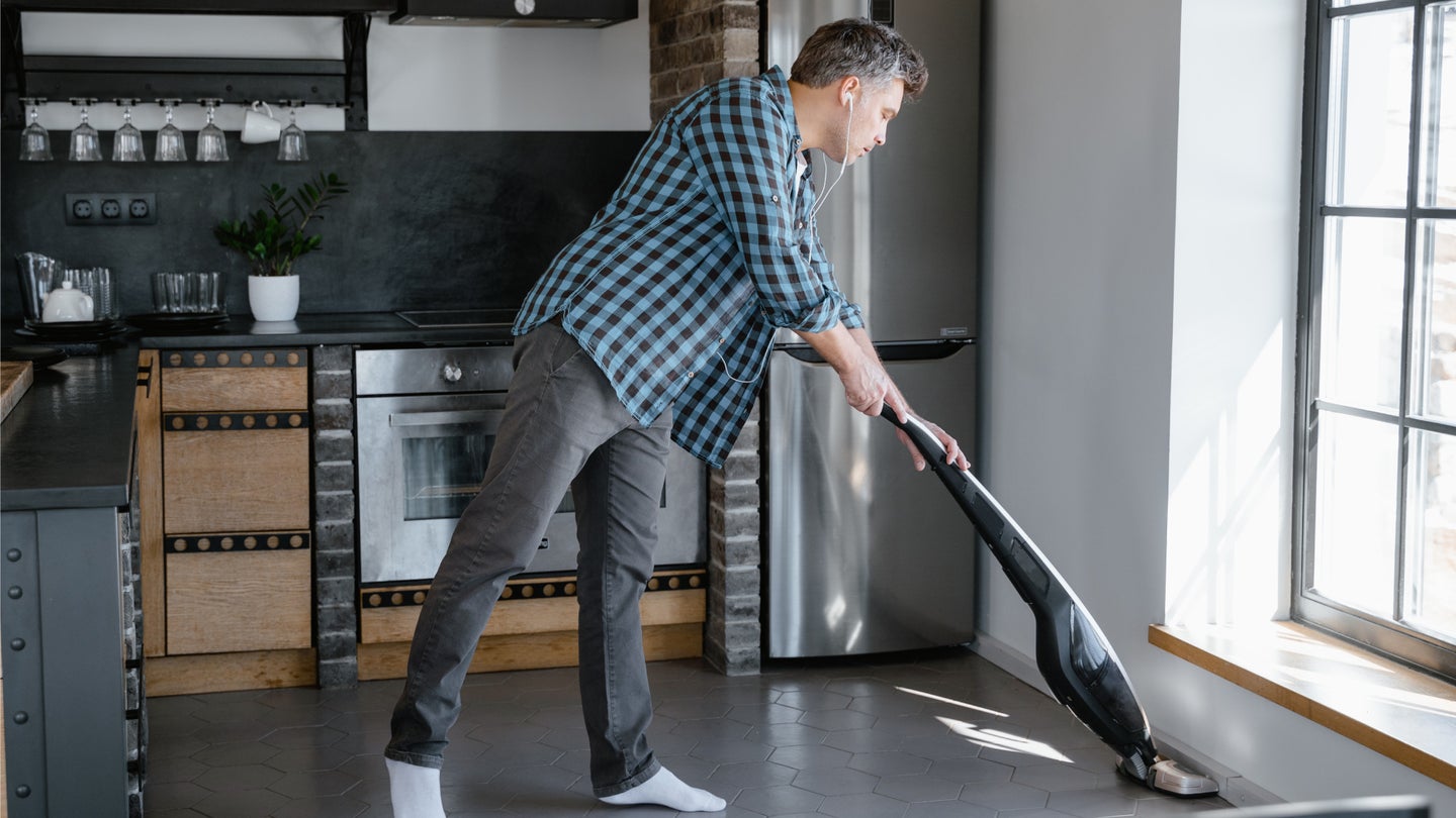 A man in a blue plaid shirt, jeans, and white socks using a cordless vacuum to clean a tile kitchen floor near a large window.