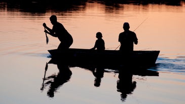 Two adults and a child in a rowboat on a calm lake at sunset. The man at the bow is paddling, the man at the back is holding a fishing rod, and the child is sitting between them.