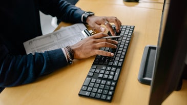 A person typing on a Windows keyboard while holding a clipboard.