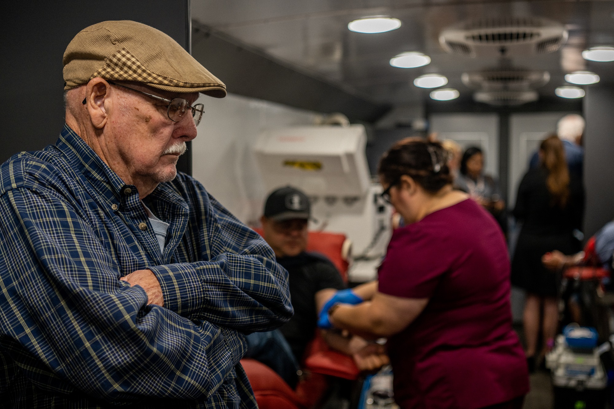 Elderly person in glasses and with mustache in blue shirt and brown cap standing in line with arms crossed at a mobile blood donor unit in Uvalde, Texas