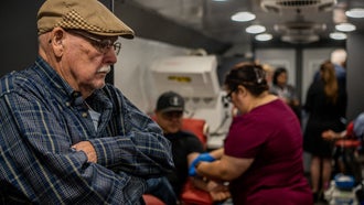 Elderly person in glasses and with mustache in blue shirt and brown cap standing in line with arms crossed at a mobile blood donor unit in Uvalde, Texas