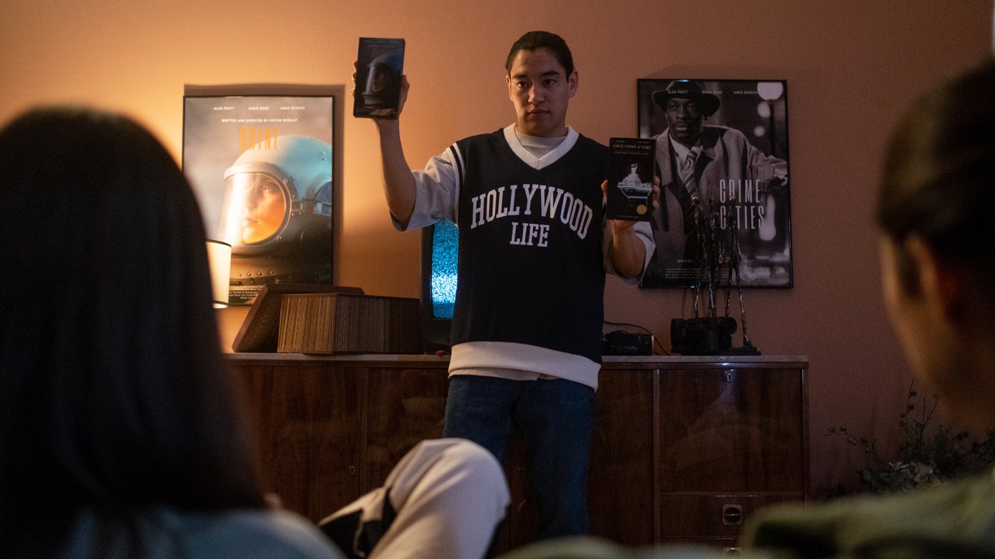 A man wearing a green-and-white jersey-style shirt that says "Hollywood Life" on it standing in front of a TV in a dim room showing two movies to two people in front of him.