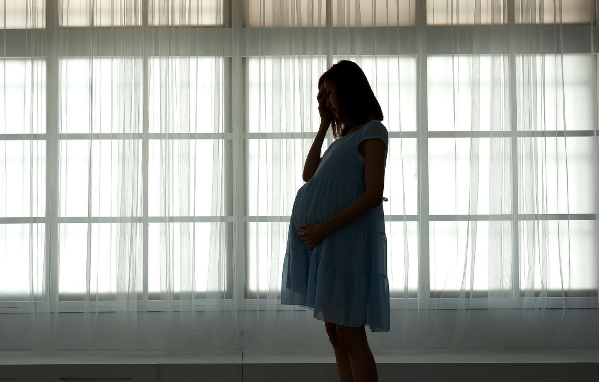 Person with long dark hair in a maternity dress in front of a window looking sad because of a miscarriage