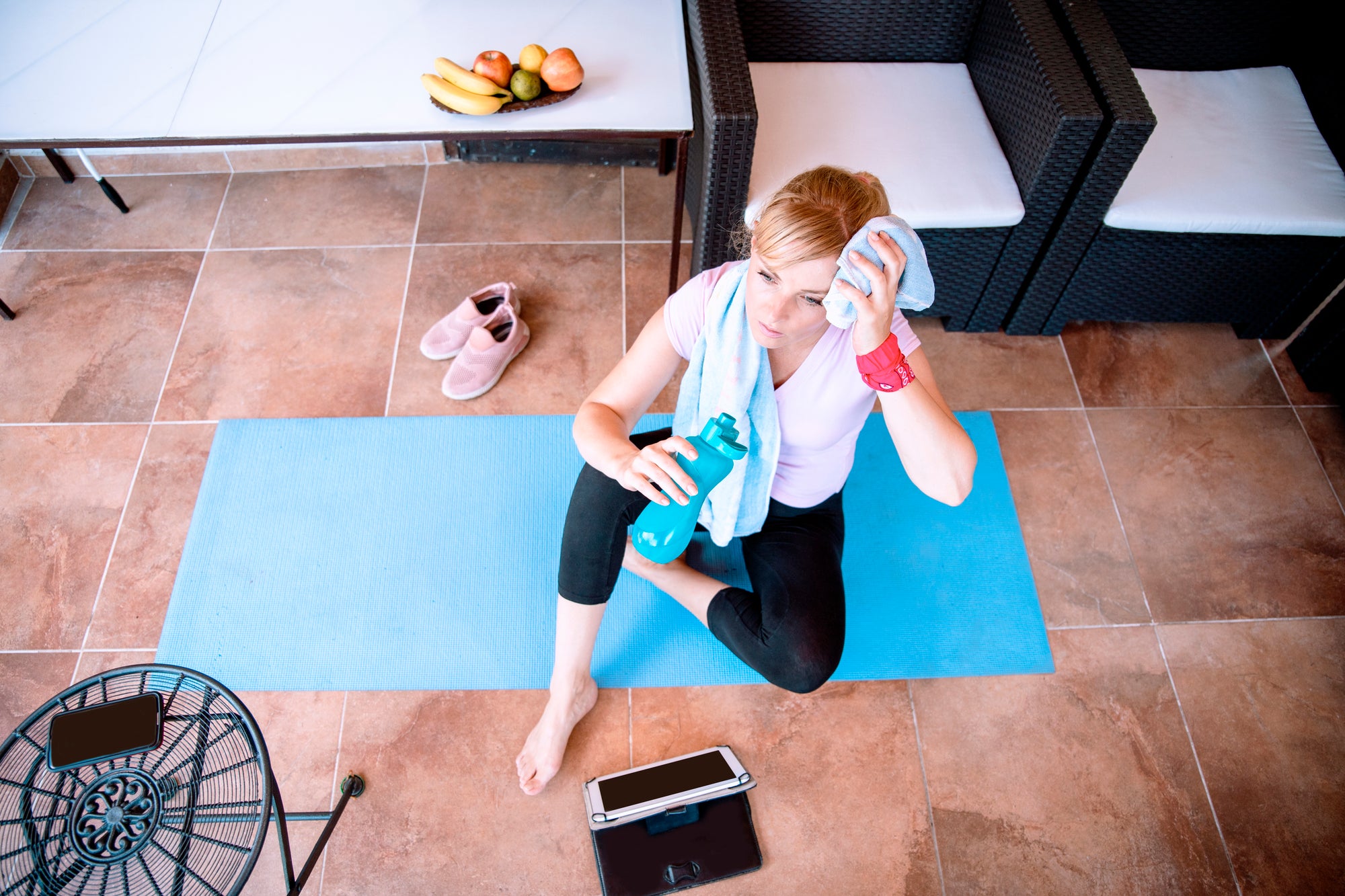Person with red ponytail in pink shirt and black workout leggings on a blue yoga mat drinking water and wiping sweat off