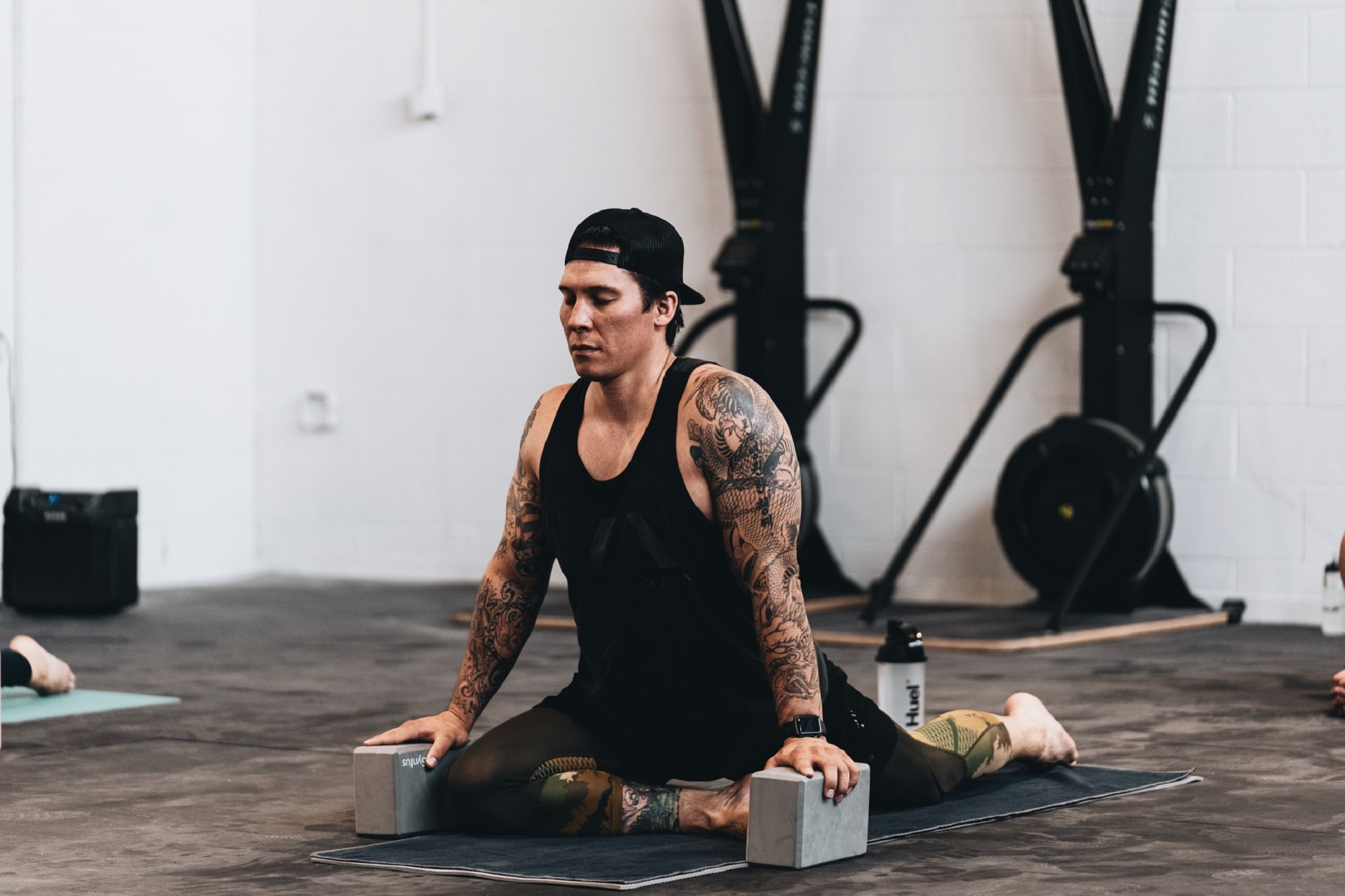 Gymgoer with tattoos and black tank top and baseball cap practicing hot yoga with blocks on a mat
