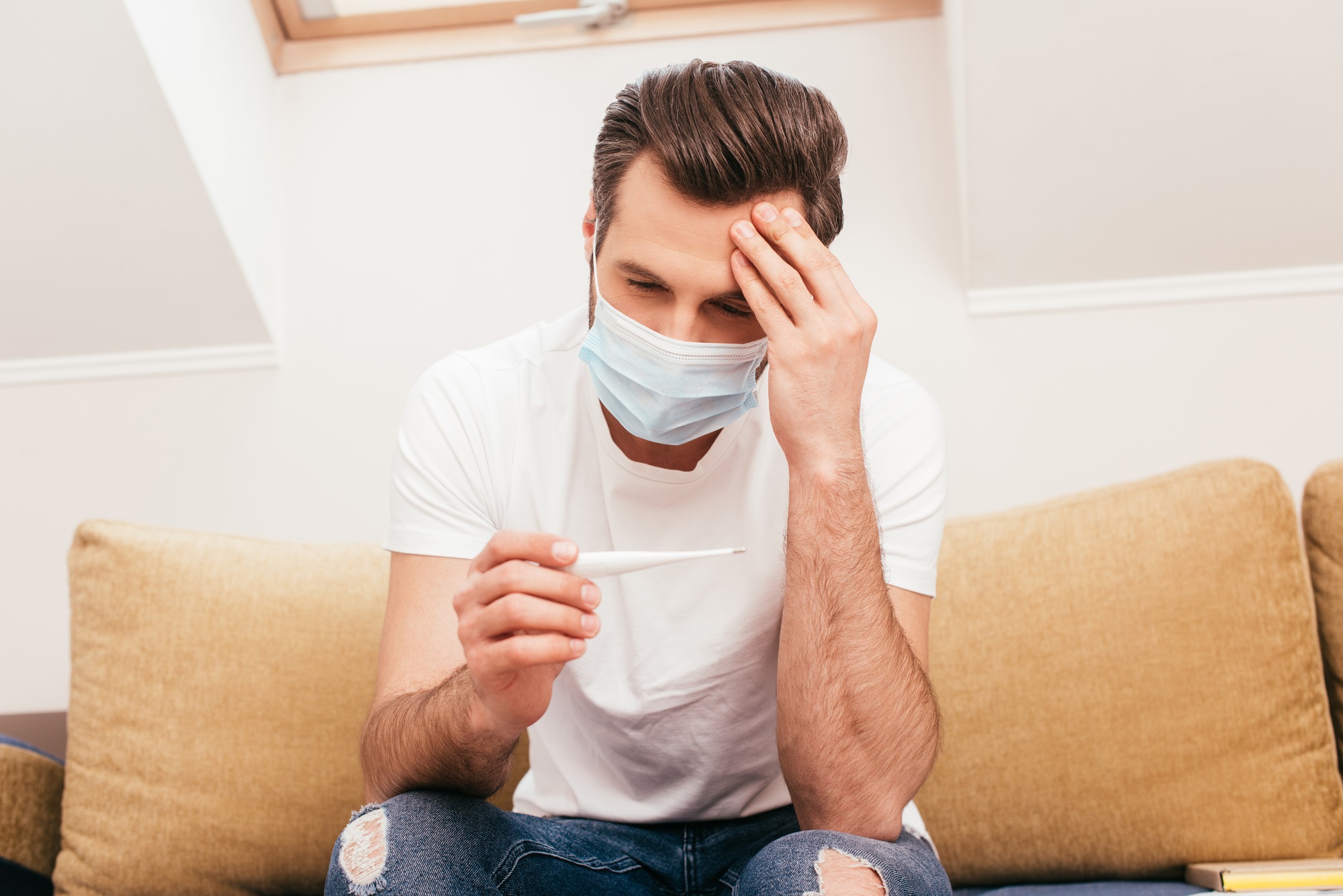 Person with short brown hair in a white t-shirt and a COVID surgical mask checking their temperature on a thermometer for COVID rebound infection