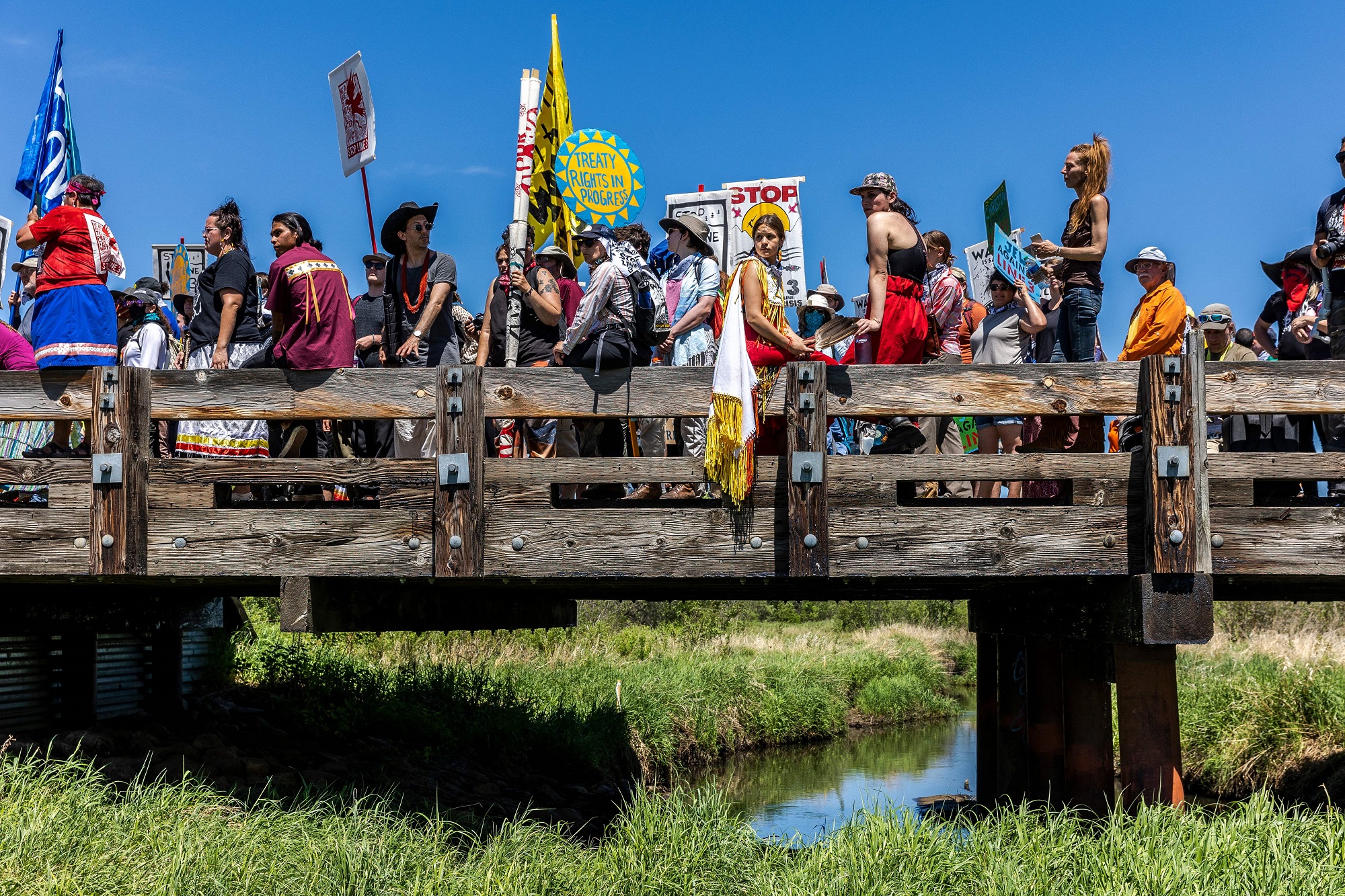 Activists in Indigenous clothing and with no pipeline signs protesting Line 3 on a bridge in Minnesota