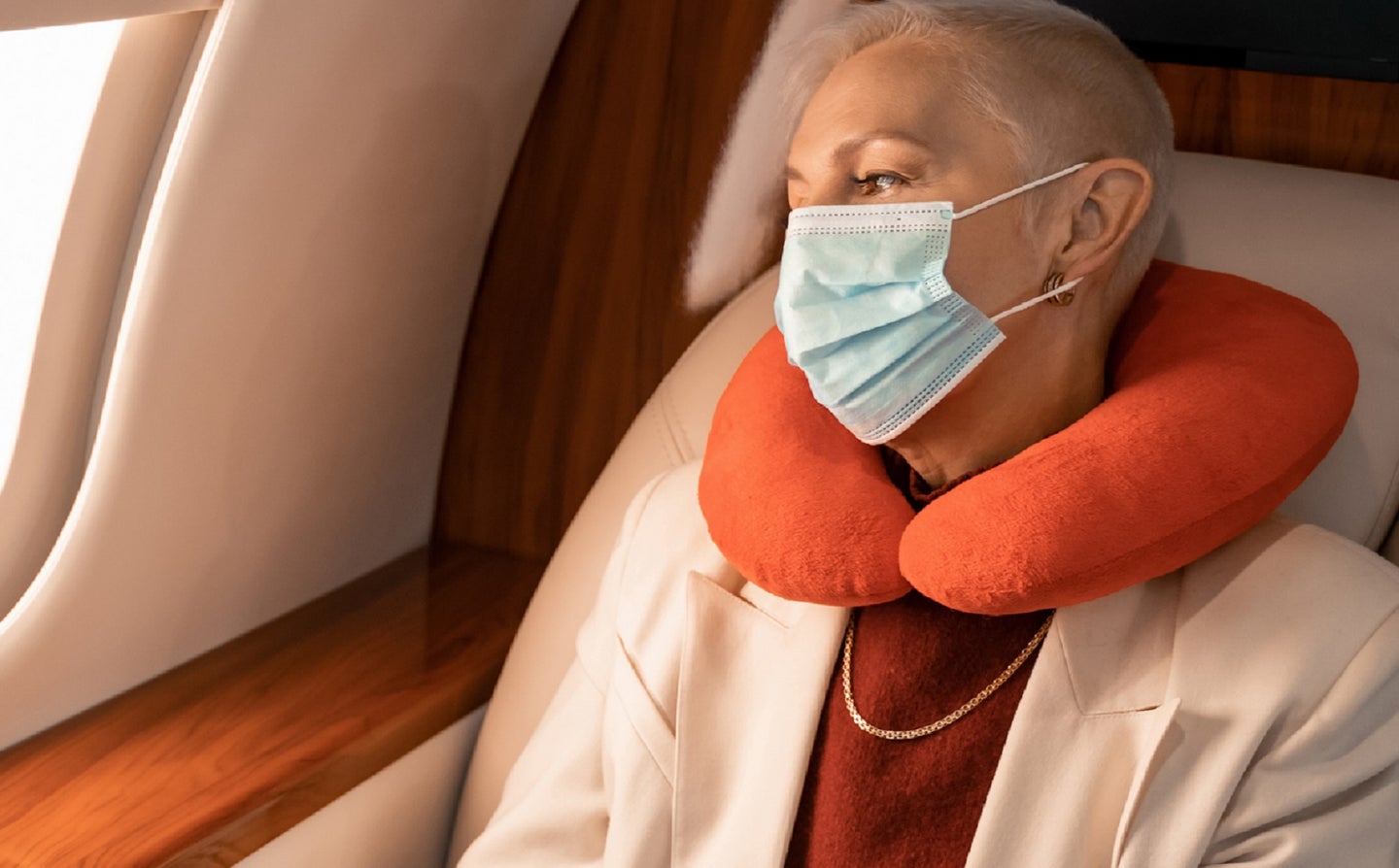 Plane traveler wearing a COVID mask with an orange neck pillow looking out window after CDC lifted COVID testing policy for international flights