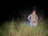 Ecologist in camo with an insect net standing at the edge of a grassy bog at night