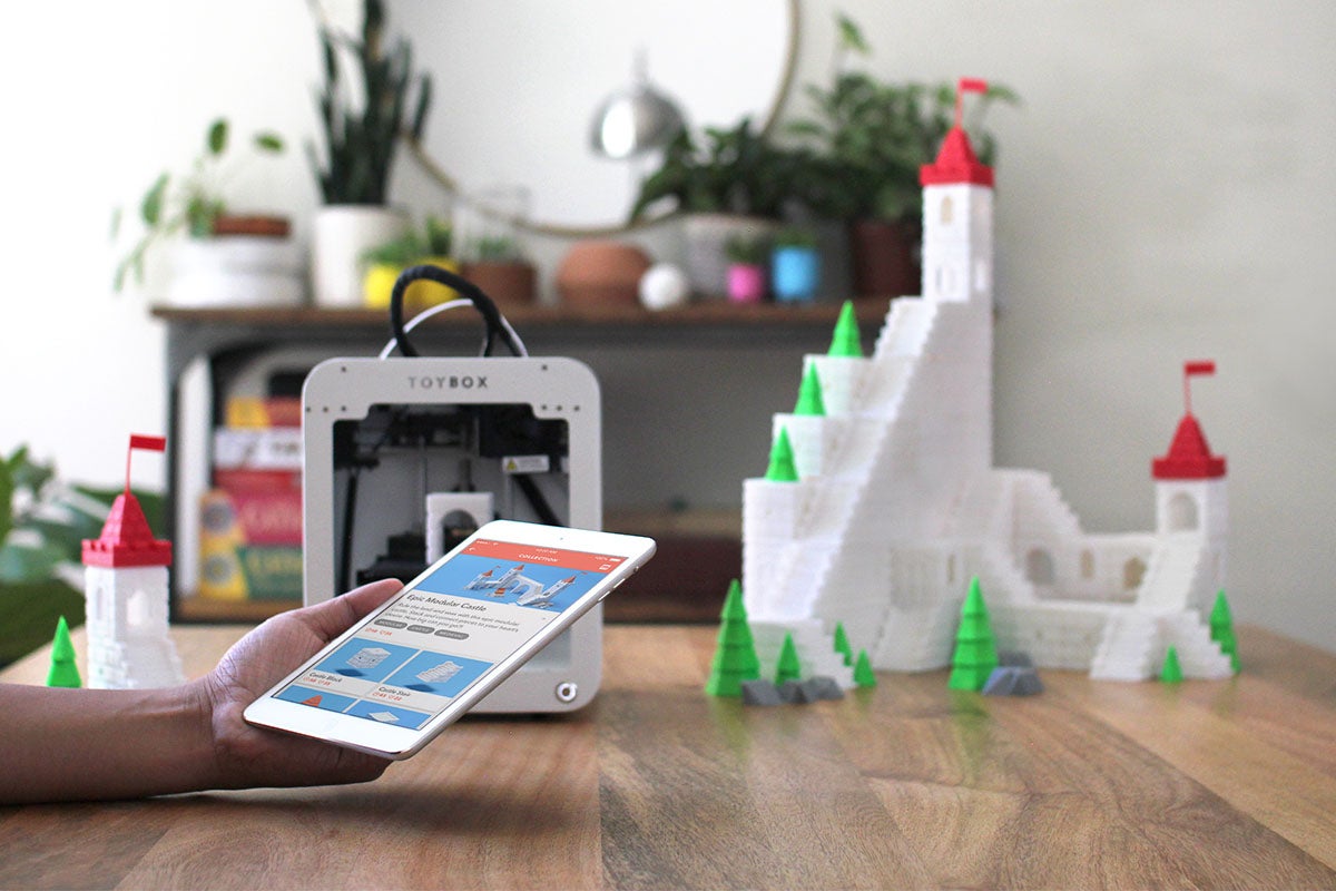 A person holding a tablet in their living room surrounded by 3D printed objects.
