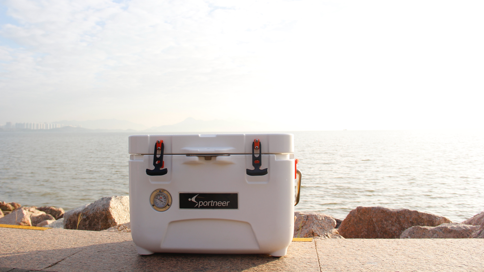 A white cooler sitting in the sun on a concrete sidewalk by a large body of water.