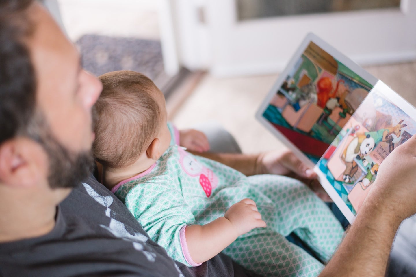Bearded dad holding a newborn baby in a blue onesie and reading a picture book