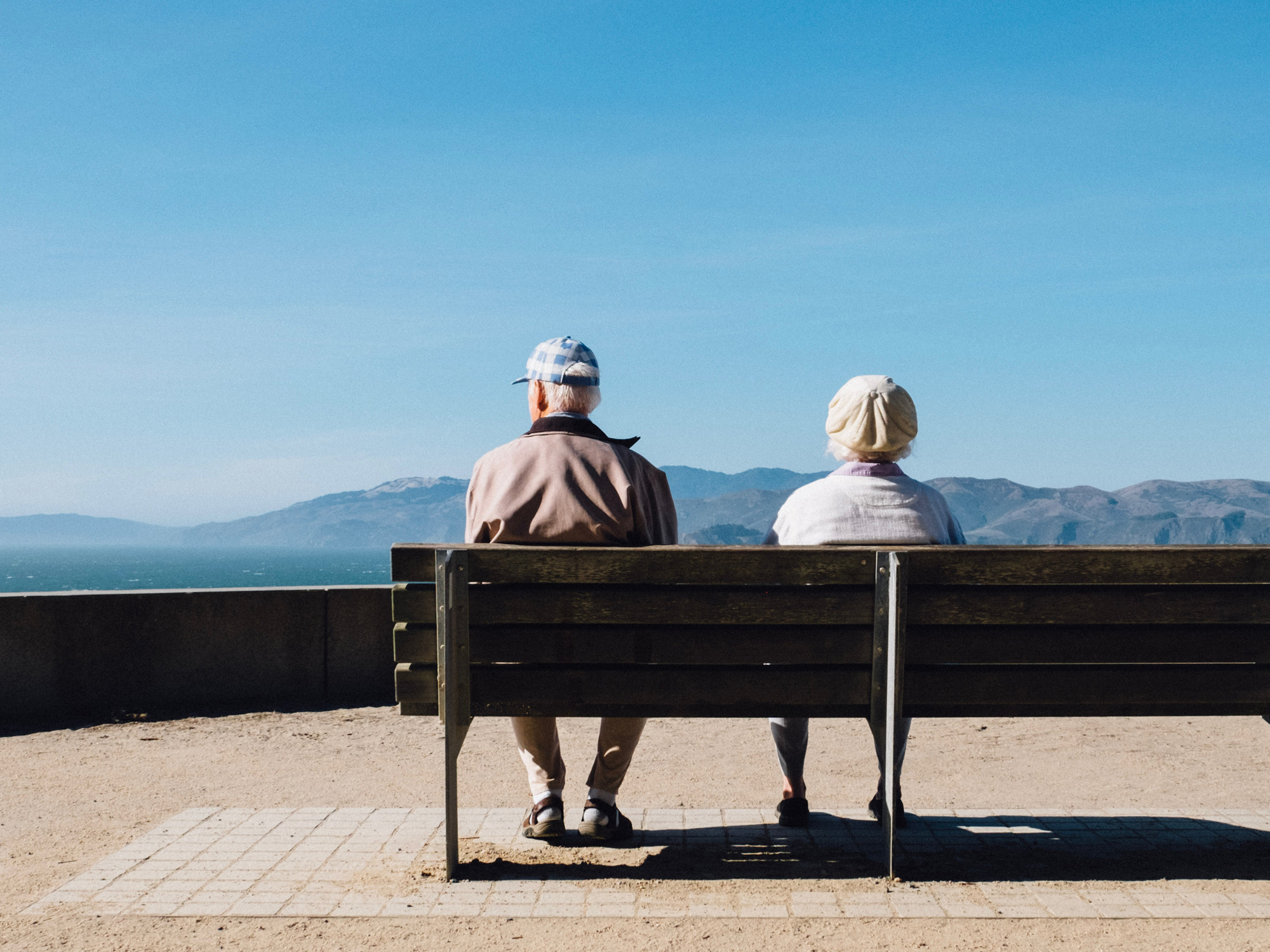 An elderly man and woman sitting on a bench on a scenic overlook in some mountains on a sunny, cloudless day.