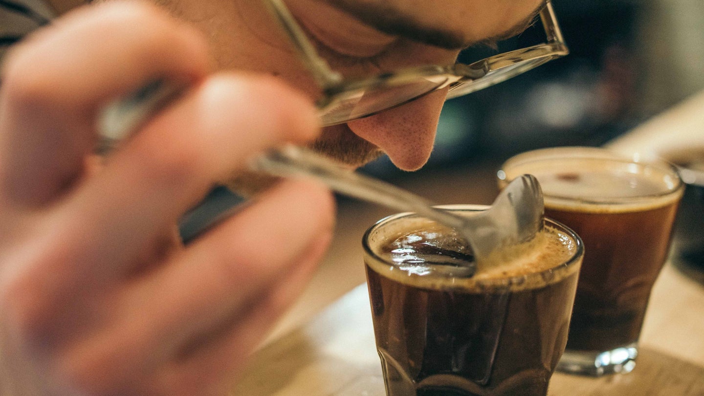 A man with a mustache and glasses taking a deep whiff of a coffee drink in front of his nose.