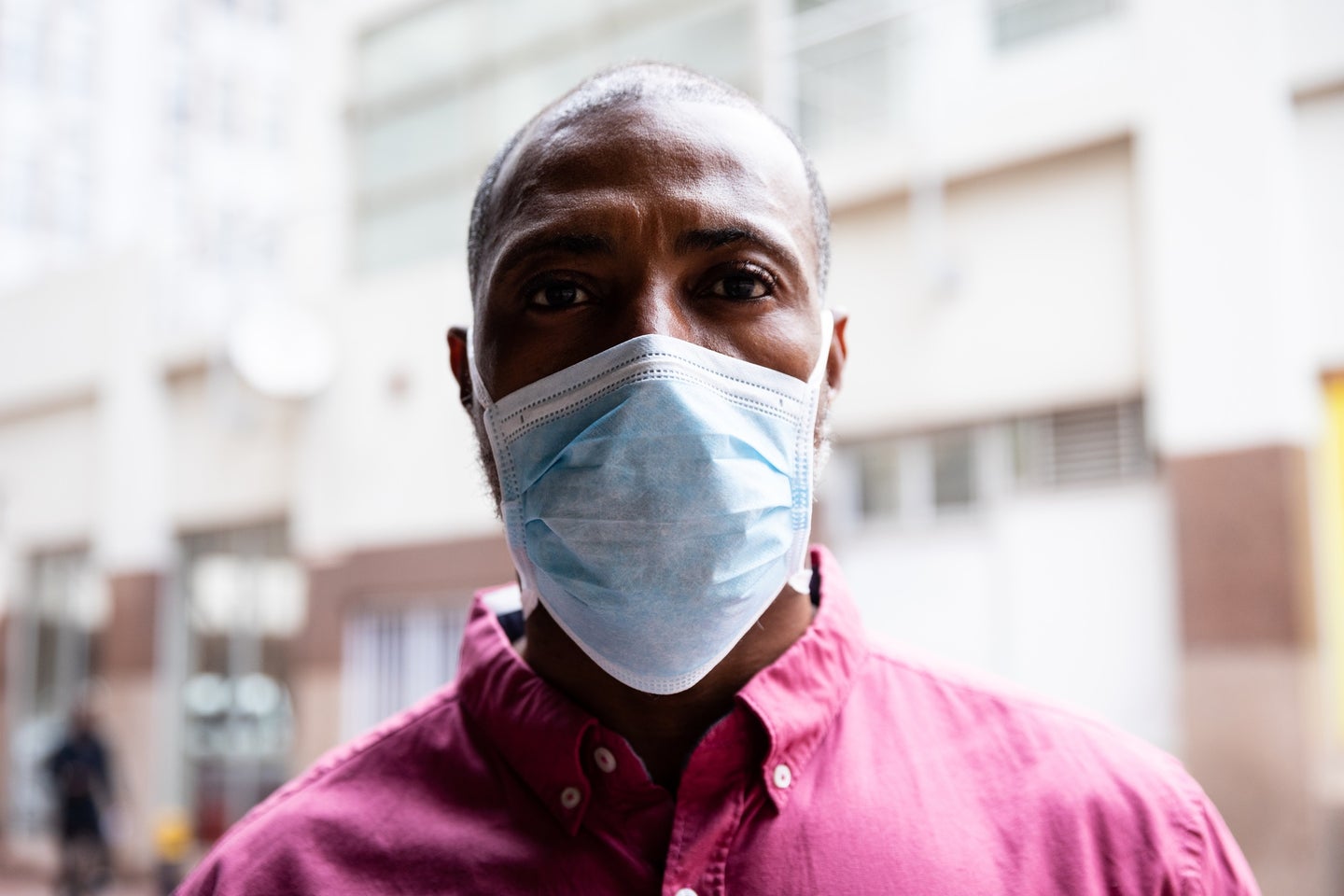 Black person with short gray hair in a red collared shirt wearing a COVID mask looking directly into the camera