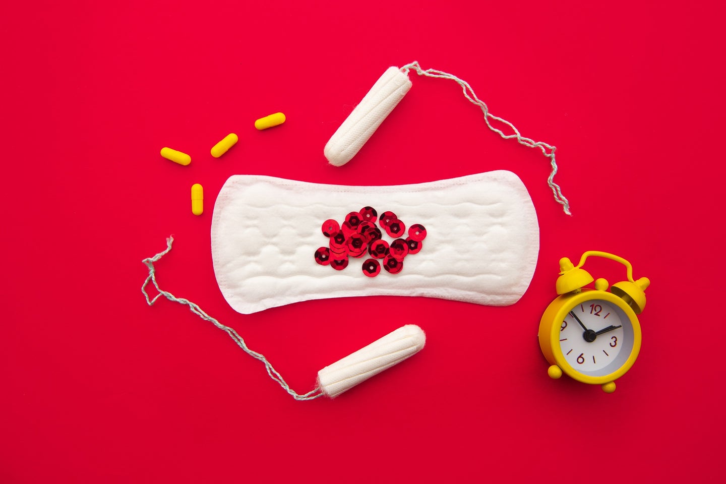 Pad with red sequins next to two tampons, yellow hormone pills, and an alarm clock to represent irregular periods