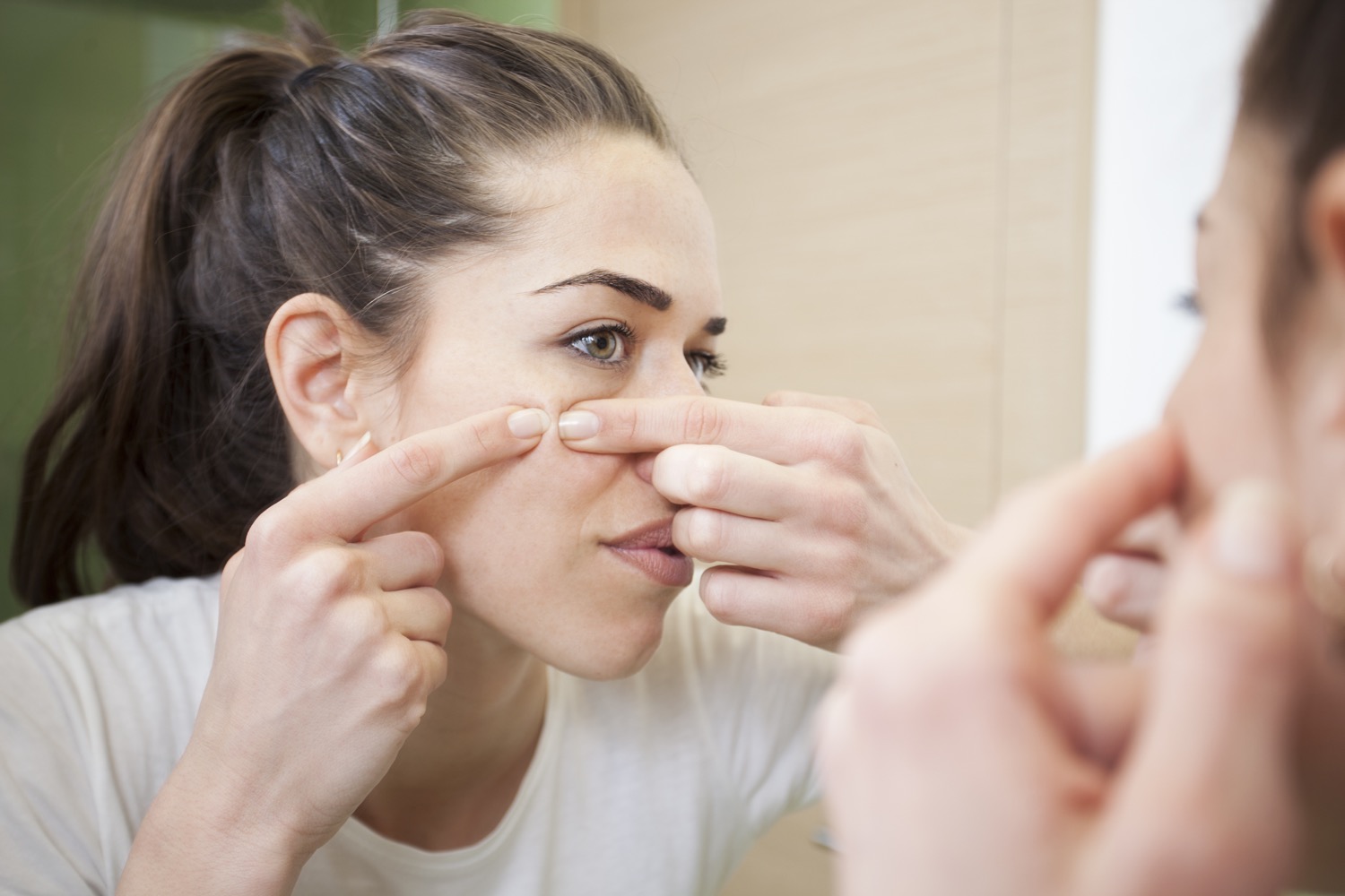 a woman looks in the mirror as she pinches a pimple on her check with her two pointer fingers