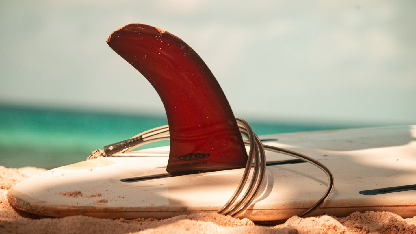 A white surfboard on beige sand, upside down, with its red fin pointing skyward.